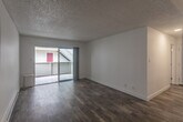 This single-floor, one-bedroom unit offers a practical and well-organized layout across a compact footprint, comprising a combined kitchen and living room, one bedroom, and a hallway. The living area is bright and open, featuring warm hardwood-style flooring and crisp white walls that contribute to a clean, minimalist aesthetic. A textured ceiling adds subtle architectural character throughout. A large sliding glass door floods the living space with natural light and provides direct access to a private outdoor balcony, offering views of the surrounding neighborhood. The kitchen is efficiently designed with white shaker-style cabinetry, light quartz countertops, and stainless steel appliances, including a refrigerator, dishwasher, and range. Light tile flooring complements the cabinetry and countertops, maintaining a cohesive, modern look. A built-in pass-through shelf connects the kitchen area to the living room, adding a functional architectural detail. The bedroom is an intimate, quiet space with the same hardwood-style flooring and neutral white walls, offering a clean canvas. A baseboard heating unit is present in the living area, ensuring year-round comfort. The hallway connects the rooms efficiently, completing this straightforward and functional residential layout.