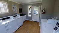 This single-floor communal laundry facility offers a clean, functional environment designed for convenient everyday use. The room is finished with warm wood-look flooring and gray-painted walls accented by crisp white crown molding, giving the space a tidy, well-kept appearance. Multiple white top-load washers and dryers are arranged along the walls, providing ample capacity for residents. Each machine features a modern payment or control panel, ensuring ease of use. A centrally located white panel door with a full-length glass insert serves as the main entrance, flooding the interior with natural light and offering a view of the surrounding parking area and landscaping. A fire extinguisher is mounted near the door, reflecting attention to safety standards. Informational signage, including a community map and notices, is displayed on the wall, keeping residents informed. The space is efficiently laid out, allowing comfortable movement between machines. Overall, the laundry facility strikes a balance between practicality and comfort, offering a bright, organized, and accessible shared amenity.