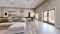 Located in Glendale, Arizona, this single-floor space offers a well-designed layout that blends modern aesthetics with comfortable, functional living. The expansive living room serves as the heart of the space, featuring vaulted ceilings with recessed lighting, wide-plank wood-look flooring, and a striking shiplap accent wall housing a wall-mounted flat-screen TV and a sleek linear electric fireplace. The room is generously proportioned, providing ample space for a large sectional sofa, a concrete-top coffee table, and a full-size pool table, making it ideal for both relaxation and entertainment. A wooden shuffleboard table lines one wall, accompanied by a curated gallery of vibrant Arizona-themed travel posters — including Sedona, Arizona, and Grand Canyon prints — adding a distinct regional character to the space. The kitchen is efficiently designed with soft gray cabinetry, white quartz countertops, and stainless steel appliances including a range and a French door refrigerator. A connecting hallway with a round mirror accent provides a smooth transition between areas. Throughout, the neutral color palette, cohesive flooring, and thoughtful décor create a warm, inviting, and polished atmosphere.