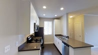 This single-floor space showcases a clean, contemporary design with a well-appointed galley-style kitchen as its centerpiece. The kitchen features crisp white cabinetry that extends to the ceiling, offering generous storage capacity on both sides of the corridor layout. Dark, polished countertops provide a striking contrast against the white cabinets, creating a modern, high-contrast aesthetic throughout the cooking space. A full suite of appliances is included, featuring a stainless steel refrigerator, an electric range with a built-in microwave above, and a dishwasher seamlessly integrated beneath the countertop. A double-basin stainless steel sink with a window above allows for natural light to filter into the kitchen, brightening the space during the day. Recessed lighting installed across the ceiling ensures the kitchen remains well-lit at all times. Dark hardwood-style flooring runs throughout, adding warmth and depth to the modern palette. A glimpse into the adjacent area reveals additional doors and hallway space, suggesting a functional and efficiently organized floor plan. The overall ambiance is polished, practical, and move-in ready, designed to appeal to those who value a clean, contemporary living environment.