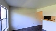 This single-floor space presents a straightforward and functional layout designed for comfortable everyday living. The main living area is defined by smooth white walls and dark flooring that provides a grounding contrast to the bright, airy atmosphere above. The open layout allows for flexible furniture arrangement and a sense of spaciousness throughout. A partial wall or breakfast bar visually connects the living area to an adjacent kitchen space, offering both separation and accessibility between the two zones. The kitchen area features warm-toned walls, creating a subtle distinction from the cooler, neutral palette of the main living space. Electrical outlets are thoughtfully positioned along the baseboards, providing practical placement for everyday use. The smooth, light-colored ceiling contributes to the overall sense of openness within the space. Natural light filters into the room, enhancing the clean, uncluttered aesthetic. The minimalist design of the space provides a blank canvas, making it highly adaptable to a variety of interior styles and furnishing choices. Overall, the space is characterized by its simplicity, practicality, and open feel.