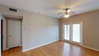This single-floor space presents a clean, open living area designed with simplicity and functionality in mind. The room features smooth hardwood-style flooring in a warm, light tone that enhances the bright and airy atmosphere throughout the space. Neutral beige walls provide a versatile backdrop, making the area adaptable to a variety of décor styles.

A ceiling fan with an integrated light fixture is centrally positioned, offering both practical air circulation and supplemental lighting. The fan's classic brass and wood finish adds a subtle traditional touch to the otherwise understated interior.

A standout feature of the space is the set of French doors along the far wall, fitted with white blinds that allow for adjustable natural light. These doors not only flood the room with daylight but also provide access to an outdoor area, contributing to the overall sense of openness. White baseboards frame the perimeter of the room, adding a crisp, finished look. The space feels well-proportioned, clean, and move-in ready, offering a comfortable and functional environment.