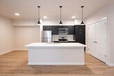 This single-floor unit showcases a modern, open-concept design with clean lines and contemporary finishes. Light-toned hardwood-style flooring runs throughout, creating a cohesive and warm aesthetic across all living spaces.

The kitchen serves as a central focal point, anchored by a generously sized island with a crisp white countertop, an undermount sink, and stylish matte black pendant lighting overhead. Dark cabinetry provides a striking contrast against the light surfaces, while stainless steel appliances — including a refrigerator, electric range, and microwave — complete the well-appointed cooking space. A double-door closet at the end of the kitchen corridor offers additional storage.

The open layout connects the kitchen seamlessly to the living area, which is bathed in natural light through large floor-to-ceiling sliding glass doors that open to a private balcony, offering outdoor views.

Two well-sized bedrooms each feature windows with horizontal blinds, allowing for natural light and exterior views. Both bathrooms are finished in neutral tones, featuring white vanities with undermount sinks, white countertops, large mirrors, and chrome fixtures. One bathroom includes a full tub/shower combo with a small accent window for added brightness.