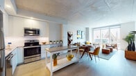 Located on Chestnut Street in Philadelphia's University City neighborhood, this single-floor residence showcases a thoughtfully designed open-concept layout with contemporary finishes throughout. The kitchen is outfitted with sleek white cabinetry, stainless steel appliances including a refrigerator, range, microwave, and dishwasher, complemented by a central island with a gray quartz countertop and light hardwood flooring. Exposed concrete ceilings lend an industrial-modern character to the open living and dining area, which is bathed in natural light from floor-to-ceiling windows. These expansive windows frame striking views of a nearby historic Gothic church and the surrounding Philadelphia skyline. The bedroom features large windows with the same captivating cityscape views, a dedicated workspace, and ample closet storage. A hallway leads to a clean, well-appointed bathroom finished with large-format tile flooring, a glass-enclosed walk-in shower, and a floating vanity. Additional closet space is accessible along the hallway corridor. The overall ambiance is bright, airy, and modern, with a neutral palette that highlights the architectural details and urban surroundings of this vibrant Philadelphia location.
