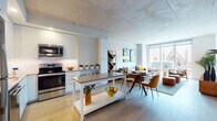 Located on Chestnut Street in Philadelphia's University City neighborhood, this single-floor residence showcases a thoughtfully designed open-concept layout with contemporary finishes throughout. The kitchen is outfitted with sleek white cabinetry, stainless steel appliances including a refrigerator, range, microwave, and dishwasher, complemented by a central island with a gray quartz countertop and light hardwood flooring. Exposed concrete ceilings lend an industrial-modern character to the open living and dining area, which is bathed in natural light from floor-to-ceiling windows. These expansive windows frame striking views of a nearby historic Gothic church and the surrounding Philadelphia skyline. The bedroom features large windows with the same captivating cityscape views, a dedicated workspace, and ample closet storage. A hallway leads to a clean, well-appointed bathroom finished with large-format tile flooring, a glass-enclosed walk-in shower, and a floating vanity. Additional closet space is accessible along the hallway corridor. The overall ambiance is bright, airy, and modern, with a neutral palette that highlights the architectural details and urban surroundings of this vibrant Philadelphia location.