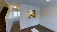 This two-floor residential space offers a functional and straightforward layout designed for comfortable everyday living. The main living area features wall-to-wall carpeting that adds warmth underfoot, while crisp white walls provide a clean, neutral backdrop throughout. A defining architectural feature of the space is the pass-through window cut into the wall between the living area and the kitchen, promoting an open feel and easy communication between the two spaces. The kitchen is equipped with white cabinetry, an electric range, and under-cabinet lighting, offering a compact yet efficient cooking environment. A long hallway extends from the living area toward a bright exterior door, allowing natural light to filter into the interior and lending a sense of depth to the floor plan. The hallway connects the various rooms of the home, contributing to a well-organized flow throughout the main level. With two floors, the space provides ample room for separate living and private sleeping quarters, making it well-suited for individuals or small households. The overall ambiance is understated and practical, with a neutral color palette and standard finishes that offer a versatile foundation for personalization.