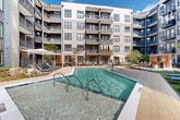 This outdoor pool amenity area in Atlanta, GA (30318) is a thoughtfully designed courtyard retreat nestled within a contemporary multi-story residential building. The centerpiece is a generously sized swimming pool with crystal-clear, blue-green water, accessible via stainless steel entry rails and framed by a wide, tiled pool deck. The surrounding courtyard is designed for both relaxation and social gatherings, featuring multiple rows of sleek, modern chaise lounges positioned to maximize sun exposure throughout the day. Shaded retreats are provided by large market umbrellas and a slatted wood pergola-style cabana structure, offering a comfortable respite from the Atlanta sun. A warm-toned wood outdoor bar or lounge station adds a social focal point to the space, complemented by stepping stone pathways that wind through manicured turf lawn areas. Decorative natural boulders and carefully selected young trees and plantings soften the hardscape and add organic texture to the environment. The courtyard is enclosed on multiple sides by the building's modern facade—characterized by horizontal lap siding in neutral tones, floor-to-ceiling windows, and private balconies—creating a sense of privacy and enclosure while framing views of the surrounding Atlanta tree canopy. The overall ambiance is that of an upscale, resort-inspired outdoor living space.