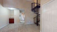 This two-floor residence offers a thoughtfully designed layout that balances open living areas with practical functionality. The main living area immediately impresses with soaring vaulted ceilings that lend a sense of grandeur, complemented by a classic white-mantled fireplace and a stylish chandelier overhead. Natural light filters in through windows with blinds, brightening the neutral-toned, carpeted floor. A striking black metal spiral staircase serves as both a functional and architectural focal point, connecting the main level to the upper floor with elegance and efficiency. The kitchen is efficiently arranged in a galley style, featuring rich dark-wood cabinetry, granite-look countertops, a double stainless steel sink, and a suite of black appliances including a refrigerator, electric range, and microwave. Hardwood-style flooring runs through the kitchen and bathroom areas, providing a clean contrast to the plush carpeting found throughout the living spaces and bedrooms. The upper level includes a versatile loft-style space with an additional sink and cabinetry, suitable for use as a study or secondary living area. The bathroom is well-appointed with a wide vanity, rich wood cabinetry, a large mirror, and a full tub-shower combination. Carpeted bedrooms feature ceiling light fixtures and ample wall space, creating comfortable, private retreats.