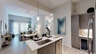 This single-floor residence showcases a contemporary open-concept design with a neutral palette of soft grays and warm whites throughout. The kitchen serves as a central highlight, featuring wood-toned cabinetry, a full suite of stainless steel appliances including a French-door refrigerator, a tile backsplash, and an island with white quartz countertops, an undermount sink, and pendant lighting overhead. The kitchen flows seamlessly into a combined dining and living area, where French doors invite natural light and connect the interior to the outdoors.

The living room offers a comfortable, well-proportioned layout with light wood-style flooring and an elevated, airy ambiance. Two bedrooms are finished with plush carpeting and ceiling fans, each providing ample wall space and access to adjacent storage. The primary bathroom features a double-sink vanity with wood-toned cabinetry, quartz countertops, and a walk-in closet. A second full bathroom includes a soaking tub and shower combination with large-format tile surround and convenient access to additional closet space. Throughout the residence, consistent finishes and thoughtful details create a cohesive, modern aesthetic.