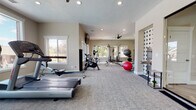 This fitness center is a comprehensive, single-floor facility designed to support a wide range of workout routines. The space is finished with neutral-toned walls and durable carpeted flooring throughout, creating a clean and functional environment. Recessed lighting and multiple ceiling fans ensure the area remains well-lit and comfortable during exercise.

The cardio zone is generously appointed with a row of treadmills, elliptical trainers, a stationary bike, and a rowing machine, all positioned along large windows that frame views of the surrounding greenery, providing an energizing workout environment. Wall-mounted televisions are conveniently placed for entertainment during cardio sessions.

The strength training area features a multi-station cable machine, a weight bench, a dumbbell rack, and an assortment of medicine balls and stability balls in various sizes, offering versatile options for resistance and functional training. A dedicated section with floor-to-ceiling mirrors supports form-focused workouts and stretching routines.

The entrance to the fitness center is accessible through a sleek glass door with black hardware, connecting to an adjacent lobby area with elegant herringbone-patterned flooring and a chandelier. A decorative cross-pattern accent wall adds a modern design touch to the interior, contributing to the overall polished and inviting atmosphere of this well-rounded fitness facility.