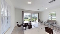This single-floor communal space is designed with a clean, modern aesthetic, characterized by crisp white walls, smooth ceilings, and light wood-look flooring that create a bright and airy atmosphere throughout. The space is divided into two interconnected areas, each serving a distinct social function while maintaining a cohesive design language.

The primary gathering area features multiple high-top bar tables paired with matching black metal stools, offering casual seating for groups. A wall-mounted flat-screen television is positioned for easy viewing, making the space well-suited for both social gatherings and informal meetings. Natural light filters in through several windows, complementing the flush ceiling fixtures that provide consistent overhead illumination.

An adjoining lounge area offers a more relaxed setting, furnished with wicker-style chairs and low tables arranged around large sliding glass doors. These floor-to-ceiling panels open to views of a landscaped garden area beyond, allowing natural light to flood the interior while providing a seamless visual connection to the outdoors. Sheer curtains frame the sliding doors, adding a subtle softness to the otherwise minimalist design. A French door in the main area provides an additional point of entry, further enhancing the space's accessibility and openness.