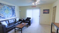This single-floor residence centers around a well-appointed living room designed for comfort and functionality. The space is anchored by a large sliding glass door fitted with vertical blinds, allowing natural light to fill the room while providing access to the outdoors. A ceiling fan with integrated lighting is mounted overhead, offering both air circulation and ambient illumination.

The living room features wall-to-wall carpeting in a neutral tone, contributing to a warm and inviting atmosphere. Seating is arranged around a rectangular wood-and-metal coffee table, creating a practical and cohesive layout. The walls are painted in a soft, light hue that enhances the brightness of the space.

Decorative elements include wall-mounted artwork that adds personality and visual interest to the room. A floor lamp in the corner supplements the overhead lighting, providing additional flexibility for illuminating the space. The sliding glass door setup suggests potential outdoor access, adding to the overall appeal of the living area. The single-floor layout offers convenient and straightforward flow throughout the residence.