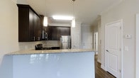 The Autumn floor plan is a one-bedroom, one-bathroom single-story residence that includes a private patio, offering an accessible and well-appointed living space. The kitchen serves as a central highlight, showcasing rich dark cabinetry that contrasts elegantly with light granite countertops. A peninsula counter extends outward, providing additional prep space and a casual bar-style seating area. Stainless steel appliances, including a gas range, microwave, and refrigerator, lend a modern, functional aesthetic. Pendant lighting above the peninsula adds focused illumination and a polished design touch, complemented by crown molding that runs along the upper perimeter of the space, adding a refined architectural detail. The flooring throughout features dark hardwood-style planks that ground the open layout with warmth and continuity. Natural light filters through the space, contributing to an inviting atmosphere. The entry foyer is visible from the kitchen, framed by a classic paneled door design. The overall aesthetic balances contemporary finishes with traditional architectural accents, creating a cohesive and comfortable living environment. The private patio extends the living space outdoors.