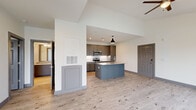 This well-designed 2-bedroom, 1-bathroom single-floor unit offers a clean, modern aesthetic with consistent light wood-look flooring and crisp white walls throughout. The open-concept living and kitchen area serves as the heart of the home, anchored by a striking blue-gray center island topped with granite countertops and a built-in sink. The kitchen is outfitted with taupe flat-panel cabinetry, a white subway tile backsplash, and a full suite of stainless steel appliances, including a refrigerator, range, and microwave. A pendant light and recessed lighting illuminate the space, complemented by a ceiling fan in the adjoining living area. The spacious primary bedroom is generously sized, featuring multiple windows that invite natural light and offer exterior views, along with a ceiling fan for added comfort. The second bedroom is similarly bright, with its own window and ceiling fan. Six-panel gray doors are used consistently throughout, adding a cohesive design detail. The bathroom is finished with a granite-topped vanity, a glass-enclosed walk-in shower with tile surround and open shelving, and a tall gray storage cabinet. A hallway connects the bedrooms and provides access to an in-unit laundry area, adding everyday convenience to this thoughtfully laid-out home.