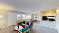 Located at Lakeview Gardens in Natick, MA, this single-floor residence offers a comfortable and functional layout with a neutral, light-filled aesthetic throughout. The open-concept living and dining area is generously sized, featuring soft wall-to-wall carpeting, light gray walls, and a textured ceiling. A bank of windows along one wall allows natural light to fill the space while providing views of the surrounding community. A wall-mounted air conditioning unit and baseboard heating ensure year-round comfort.

The living area transitions seamlessly into a dedicated dining space, accommodating a small table and chairs. Adjacent to the dining area, the kitchen features granite countertops, white cabinetry, a black refrigerator, an electric range, and a sink with a pass-through counter that connects to the main living space.

The bedroom is carpeted and well-proportioned, with windows that bring in natural light and a dedicated closet offering ample hanging and shelving space. The full bathroom is finished with classic white hexagonal tile flooring, white subway tile wainscoting, a wall-mounted sink, a mirrored medicine cabinet, and a shower-tub combination, creating a clean and timeless look.