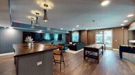 This thoughtfully designed community amenity space combines entertainment, comfort, and modern style into a single, open-concept floor. Rich hardwood flooring flows throughout, anchoring the warm, contemporary aesthetic defined by textured brown wall panels and bold teal accent wallcoverings.

The wet bar area serves as a central gathering point, featuring a curved island with a solid wood countertop, mid-century modern bar stools, pendant lighting with sleek metallic fixtures, a beverage refrigerator, and slatted wood wall accents. Adjacent to the bar, a large wall-mounted display — spanning the width of the room — provides an ideal setup for communal viewing, complemented by teal upholstered accent chairs.

The lounge area is anchored by an oversized sectional sofa and a round wood coffee table, bathed in natural light from floor-to-ceiling windows and glass doors that open to an outdoor balcony with views of the surrounding community. A dedicated game zone features a full-size shuffleboard table beneath a linear pendant light, with additional game tables visible through interior glass partitions. The space is accessible via double doors and includes restroom facilities, making it a fully self-contained amenity suited for social gatherings and everyday recreation.