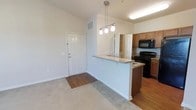 This single-floor space features an open-concept design that integrates the living area and kitchen into one cohesive layout. The living area is finished with plush carpeting, while the kitchen and entryway transition to warm hardwood-style flooring, creating a natural visual distinction between spaces. A peninsula counter with a light stone surface serves as the centerpiece of the kitchen zone, offering additional prep space and casual seating potential. Three pendant lights hang above the peninsula, providing focused task lighting while contributing to the overall aesthetic. The kitchen is equipped with dark black appliances, including a range and over-the-range microwave, complemented by medium-toned wood cabinetry that adds warmth to the space. The entryway, marked by a classic white paneled door, offers a clean and welcoming entrance. Neutral wall tones throughout create a bright, airy atmosphere that enhances the sense of openness. Elevated ceilings with visible HVAC venting contribute to a spacious feel. The layout is functional and efficient, making it well-suited for everyday living.