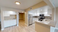 This single-floor residence offers a clean, modern aesthetic with thoughtful finishes throughout. The centerpiece of the home is a well-designed kitchen that immediately draws the eye with its crisp white cabinetry extending to the ceiling, providing generous storage space. A tasteful mosaic tile backsplash in neutral earth tones adds texture and visual interest, complementing the light granite countertops that offer ample prep space. A full suite of stainless steel appliances — including a refrigerator, range with electric cooktop, over-the-range microwave, and dishwasher — gives the kitchen a polished, contemporary feel. The kitchen opens to the rest of the living areas through a wide pass-through, creating an open and connected layout. Light-toned large-format tile flooring runs throughout, lending a sense of continuity and easy maintenance. Recessed lighting in the kitchen provides bright, even illumination, while additional ceiling fixtures light the connecting hallway and adjacent rooms. The warm two-tone wall color — soft white and muted gold — adds warmth to the interior without overwhelming the neutral palette. A secondary sink area is also visible, suggesting a dedicated laundry or utility space nearby. Overall, the space presents a clean, move-in-ready environment with modern upgrades throughout.