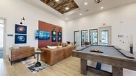 Located in Midlothian, TX, this well-appointed single-floor community clubhouse offers a generously sized, open-concept layout designed for both socializing and productivity. The main lounge area serves as the heart of the space, boasting soaring ceilings with a distinctive reclaimed wood tray ceiling inset and recessed lighting. Light wood-look tile flooring flows throughout, complementing the warm, modern rustic aesthetic. A full-size billiards table anchors the game area, surrounded by comfortable leather seating, accent chairs, and a bold terracotta-red feature wall adorned with billiard ball-themed artwork. Large black-framed glass doors allow natural light to pour in while offering views to the exterior. The kitchen area is equally impressive, featuring dark gray cabinetry, a striking patterned tile backsplash, sleek white quartz countertops, and a large island with a herringbone brick base — ideal for gatherings and catered events. A private conference room provides a quieter setting, appointed with plush carpet, a large white table, burnt orange seating, tall windows overlooking greenery, and a herringbone wood accent wall. Together, these areas create a versatile, design-forward space suited for community events, casual recreation, and professional use.
