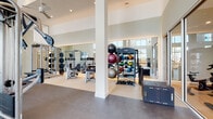 Located at 6850 Foxtail Palm Way in Bradenton, Florida, this well-appointed fitness center offers a spacious, single-floor layout designed to support a wide variety of workout routines. The facility is characterized by soaring ceilings with warm wood beam accents and elegant pendant lighting, creating an open and inviting atmosphere throughout.

Floor-to-ceiling windows line the cardio zone, allowing ample natural light to pour in while offering views of swaying palm trees outside — an uplifting backdrop for treadmills, ellipticals, and a stair climber. The durable dual-tone flooring transitions seamlessly between zones, delineating cardio and strength areas with practical elegance.

The strength training section is comprehensively equipped with a multi-station cable machine, a full rack of Cybex dumbbells, resistance machines, and a diverse collection of medicine balls, stability balls, and kettlebells. Large wall-to-wall mirrors expand the sense of space and allow users to monitor their form throughout their session.

A rowing machine and plyo box further support functional and high-intensity training needs. The facility is accessed through sleek glass doors framed by a minimalist entryway with warm wood cabinetry and tasteful greenery. Clean white walls and recessed lighting throughout contribute to the polished, modern aesthetic of this thoughtfully designed fitness space.
