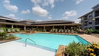 This community amenity space is thoughtfully designed to offer both relaxation and recreation. At its center is a generously sized resort-style swimming pool with crystal-clear turquoise water, framed by a stone pool deck lined with cushioned lounge chairs. The adjacent clubhouse building features a low-pitched roof with wide overhanging eaves, creating a covered outdoor corridor that connects the interior spaces to the pool area.

Inside, the clubhouse is defined by soaring ceilings accented with bold dark geometric beam detailing and recessed lighting throughout. The main lounge area is an open, airy space furnished with patterned accent chairs and round tables, anchored by a wet bar with sleek dark cabinetry and bar seating. Large windows dressed with sheer floor-to-ceiling curtains flood the interior with natural light while offering views of the surrounding greenery.

A dedicated games room houses a full-size billiards table beneath a large statement pendant light, with additional seating nearby. Separate flex spaces feature bar-height tables and wall-mounted televisions, suitable for private gatherings or meetings. Striped carpeting runs throughout the interior, complementing the contemporary neutral color palette and contributing to the polished, cohesive ambiance of the entire space.