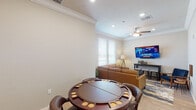 Located in Tontitown, Arkansas, this single-floor space offers a thoughtfully designed living area that blends comfort and functionality. The main room is anchored by a generous L-shaped leather sectional sofa in warm cognac tones, positioned atop a bold abstract area rug that adds a striking visual element to the light hardwood flooring beneath. A wall-mounted flat-screen TV and a narrow bar-height console table with stools provide an entertainment-ready setup, while a ceiling fan with integrated lighting ensures year-round comfort. Crown molding runs along the perimeter of the ceiling, lending a polished, refined character to the space, and recessed lighting offers clean, even illumination throughout. Natural light filters in through large windows fitted with horizontal blinds, offering views of the surrounding neighborhood. A dedicated game area features a round poker-style table with built-in cup holders and cushioned chairs, perfect for entertaining. Decorative touches such as abstract wall art, small sculptural accents, and a tripod floor lamp contribute to the room's curated, inviting ambiance. A paneled interior door connects to an adjacent room, enhancing the overall flow and accessibility of the space.