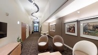 This single-floor space presents a polished and professional interior designed with both functionality and aesthetic appeal in mind. The central corridor serves as the spine of the layout, drawing the eye toward a frosted glass entry door at the far end, which allows diffused natural light to filter into the space. Overhead, a striking sculptural pendant light fixture with a cascading, wave-like design adds a dramatic focal point to the hallway, complementing the clean white ceiling above. The walls are finished in a warm taupe tone, lending a refined and welcoming atmosphere throughout. The waiting or reception area is appointed with distinctive rounded pod-style chairs in a neutral brown upholstery, arranged to encourage comfortable seating. Framed artwork depicting musical subjects adorns the walls, adding a cultural and artistic dimension to the environment. Dark-patterned carpeting runs the length of the corridor and seating area, providing visual contrast against the lighter wall tones. Along one side of the hallway, warm wood-paneled doors lead to individual private rooms, suggesting a professional services or medical office setting. The overall ambiance is contemporary yet inviting, balancing modern design elements with a calm, neutral palette.