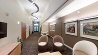 This single-floor space presents a polished and professional interior designed with both functionality and aesthetic appeal in mind. The central corridor serves as the spine of the layout, drawing the eye toward a frosted glass entry door at the far end, which allows diffused natural light to filter into the space. Overhead, a striking sculptural pendant light fixture with a cascading, wave-like design adds a dramatic focal point to the hallway, complementing the clean white ceiling above. The walls are finished in a warm taupe tone, lending a refined and welcoming atmosphere throughout. The waiting or reception area is appointed with distinctive rounded pod-style chairs in a neutral brown upholstery, arranged to encourage comfortable seating. Framed artwork depicting musical subjects adorns the walls, adding a cultural and artistic dimension to the environment. Dark-patterned carpeting runs the length of the corridor and seating area, providing visual contrast against the lighter wall tones. Along one side of the hallway, warm wood-paneled doors lead to individual private rooms, suggesting a professional services or medical office setting. The overall ambiance is contemporary yet inviting, balancing modern design elements with a calm, neutral palette.