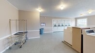 Located at 3111 S Leonard Springs Rd in Bloomington, Indiana, this shared laundry facility offers a clean, functional, and spacious environment designed to accommodate high-volume use. The single-room layout is efficiently organized, with rows of top-loading washing machines lining one side and stacked dryer units positioned along the opposite wall, providing substantial laundry capacity for residents. The speckled terrazzo-style flooring is durable and easy to maintain, while the textured popcorn ceiling adds a classic finish to the space. Warm beige walls with a white chair rail accent create a neutral, welcoming ambiance throughout. Natural light streams in through multiple white-framed windows positioned along the walls, brightening the interior and reducing the reliance on the flush-mounted ceiling light fixtures. A rolling laundry cart and a waste bin are conveniently available for users. A double door entry with a floor mat provides easy access to the facility. Informational postings are displayed on the wall for user guidance. The open floor plan allows for comfortable movement between machines, making the laundry process straightforward and efficient for all users.