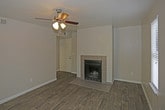 This single-floor space presents a clean, straightforward layout centered around a welcoming living area. The room is anchored by a tile-surround fireplace with a classic black insert, adding a functional and visually grounding focal point to the space. The hearth extends outward with matching tile flooring, creating a defined transition between the fireplace surround and the dark-toned hardwood flooring that spans the rest of the room.

The walls are finished in a neutral off-white tone, contributing to a bright and open atmosphere. A ceiling fan with an integrated light fixture is centrally mounted, offering both ambient lighting and air circulation. Natural light enters through a vertical window fitted with white blinds, further enhancing the airy feel of the room.

The overall design is understated and practical, with clean lines and minimal ornamentation. The combination of dark hardwood flooring, neutral walls, and the tiled fireplace creates a balanced contrast that gives the space a tidy, move-in-ready appeal. The single-floor configuration ensures convenient accessibility throughout.