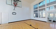 This exceptional indoor sports facility is centered around a full regulation-marked basketball court finished in gleaming hardwood, with crisp court lines clearly delineated across the polished surface. The space is defined by its soaring ceiling height, creating an open and airy atmosphere well-suited for athletic activity. A wall-mounted basketball system with a clear acrylic backboard and standard hoop is installed at one end, while a decorative plaque adorns the white wall beneath it.

Natural light pours generously into the court through an expansive, multi-pane window wall that rises dramatically toward the ceiling, offering views of lush greenery and the surrounding landscape. A set of glass double doors provides access to an adjacent area, maintaining visual connectivity throughout the facility. Large interior windows along another wall look directly into a well-equipped fitness center, visible with a full array of cardio and strength training machines, seamlessly integrating the two athletic spaces.

The clean white walls, dark baseboards, and bright hardwood floors combine to give the court a polished, professional aesthetic. The abundance of natural light, generous vertical volume, and thoughtful layout make this an exceptional private sports and wellness facility.