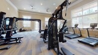 This fitness center in Port Orange, Florida, is a thoughtfully designed, single-floor facility offering a comprehensive workout environment. The space features elevated ceilings accented by crisp crown molding, recessed lighting, and ceiling fans that contribute to a bright and comfortable atmosphere. The warm, neutral-toned walls are complemented by white wainscoting panels, lending the space a polished, upscale aesthetic.

Flooring throughout consists of durable, multi-toned carpet tiles in a geometric pattern, providing both comfort and visual interest. Large windows fitted with blinds allow generous natural light to filter in, creating an inviting and energizing environment.

The cardio zone includes treadmills, elliptical cross-trainers, a StairMaster, and stationary bikes, many of which are equipped with individual monitors. A dedicated strength training area houses multiple cable and weight machines arranged along a mirrored wall, which enhances the sense of space and allows users to monitor their form. A dumbbell rack provides free weight options. A sliding frosted-glass barn door elegantly divides sections of the facility, adding a functional yet stylish design detail. The overall layout is spacious and well-organized, supporting a full range of fitness activities.