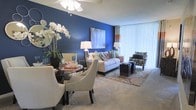 This single-floor residence showcases a thoughtfully designed open-plan layout that seamlessly integrates a dining area and living room into one cohesive, inviting space. The most striking architectural feature is a bold navy blue accent wall in the living area, which serves as a dramatic backdrop for a light-colored sofa adorned with an array of colorful decorative pillows. A patterned armchair and dark espresso-toned side tables complement the seating arrangement, while a soft, patterned area rug anchors the lounge zone. Floor-to-ceiling vertical blinds span the exterior wall, diffusing generous amounts of natural light throughout the space while offering privacy. The dining area features a sleek glass-top table surrounded by elegant cream upholstered chairs with nailhead trim detailing, paired with a striking floral arrangement for a refined aesthetic. Plush wall-to-wall carpeting runs throughout, adding warmth and comfort underfoot. A ceiling fan overhead provides practical functionality while contributing to the overall clean, modern ambiance. The space balances bold design choices with neutral tones, creating a welcoming and visually dynamic environment.
