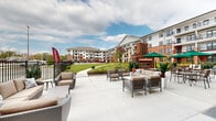 This outdoor amenity space is a thoughtfully designed courtyard patio situated at the base of a multi-story residential building featuring a classic combination of red brick and white exterior paneling. The patio is finished with large-format concrete pavers that provide a clean, modern surface throughout the generous open-air area.

The space is organized into distinct zones to accommodate a variety of outdoor activities. A lounge area offers plush, neutral-toned sectional sofas and armchairs arranged around a low coffee table, ideal for relaxed conversation. Adjacent to this is a dining area furnished with round tables and chairs, shaded by vibrant green market umbrellas that add a lively accent to the setting.

A standout architectural feature is a tall, freestanding brick fireplace and chimney structure positioned along the perimeter, serving as a functional focal point and visual anchor for the outdoor dining zone. Potted tropical plants and manicured landscaping are integrated throughout, adding greenery and a resort-like ambiance.

The courtyard opens toward a well-maintained lawn with walking paths, a pool area visible in the distance, and broad views of the surrounding community buildings. Large windows and private balconies of the building overlook the patio, reinforcing the seamless connection between interior and exterior living spaces.