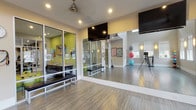 This yoga and fitness studio is thoughtfully designed to support a wide range of movement and wellness practices across a single, efficiently organized floor. The main practice area features smooth hardwood-style flooring that provides an ideal surface for yoga, stretching, and group exercise. Full-wall mirrors line the studio, allowing practitioners and instructors to monitor form and alignment with ease. Overhead, recessed lighting and a ceiling fan work in tandem to maintain a comfortable and well-lit environment during sessions. Mounted flat-screen televisions offer visual instruction or guided content for participants. A glazed partition wall with large glass panels connects the main studio to an adjacent brightly colored support area, lending the space a sense of openness and visual depth. Built-in benches provide convenient seating along the perimeter. A dedicated equipment zone houses an organized collection of stability balls in various sizes and colors, resistance bands, and exercise bars on storage racks. Natural light filters in through multiple windows fitted with roller shades, offering views to the outdoors while maintaining privacy. Instructional posters displayed on the walls add a functional, educational element to the overall design. The studio's clean, neutral palette and purposeful layout create a focused and energizing atmosphere suited for both individual and group fitness sessions.