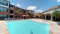 This outdoor lounge area is a beautifully appointed resort-style retreat designed for relaxation and leisure. At its centerpiece sits a generously sized swimming pool with sparkling turquoise water, offering an inviting atmosphere on sunny days. The pool deck is lined with a generous arrangement of lounge chairs, providing ample space for sunbathing and unwinding. Shaded cabanas and umbrella-covered seating areas are thoughtfully positioned along the poolside, offering comfortable retreats from the sun. Lush palm trees add a tropical touch to the landscape, enhancing the resort ambiance. The surrounding architecture features classic brick multi-story residential buildings with large windows that overlook the pool area, complemented by a charming stucco-accented clubhouse or pavilion structure with arched detailing. An ornate iron fence defines the perimeter of the lounge, adding an elegant boundary to the space. The open-air setting benefits from abundant natural light under broad blue skies, creating a bright and welcoming environment. The cohesive design of the landscaping, seating, and architectural elements come together to deliver a polished and upscale outdoor experience.