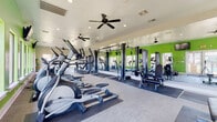 This fitness center is designed to support a comprehensive workout experience across a single, thoughtfully organized floor. The space is immediately distinguished by its bold lime-green accent walls, which energize the environment and contrast with neutral tones throughout the rest of the room. Full-length mirrors line one wall, visually expanding the space and allowing users to monitor their form during exercise.

The cardio zone is well-appointed with multiple treadmills, elliptical machines, and stationary bikes, positioned near large windows that provide views of the surrounding outdoor landscape and allow ample natural light to fill the room. Wall-mounted televisions are conveniently located in this area for entertainment during workouts.

A dedicated strength training section features cable and resistance machines alongside a tiered dumbbell rack stocked with a wide range of free weights, accompanied by an adjustable workout bench. Rubber flooring mats are placed throughout the equipment zones for safety and durability.

A separate open-floor area with light wood-style flooring serves as a functional training or stretching space, equipped with medicine balls and a yoga mat, ideal for flexibility work or bodyweight exercises. Ceiling fans and recessed lighting are installed throughout, ensuring consistent airflow and visibility. The overall design is practical, clean, and conducive to focused, effective training.