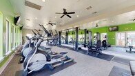 This fitness center is designed to support a comprehensive workout experience across a single, thoughtfully organized floor. The space is immediately distinguished by its bold lime-green accent walls, which energize the environment and contrast with neutral tones throughout the rest of the room. Full-length mirrors line one wall, visually expanding the space and allowing users to monitor their form during exercise.

The cardio zone is well-appointed with multiple treadmills, elliptical machines, and stationary bikes, positioned near large windows that provide views of the surrounding outdoor landscape and allow ample natural light to fill the room. Wall-mounted televisions are conveniently located in this area for entertainment during workouts.

A dedicated strength training section features cable and resistance machines alongside a tiered dumbbell rack stocked with a wide range of free weights, accompanied by an adjustable workout bench. Rubber flooring mats are placed throughout the equipment zones for safety and durability.

A separate open-floor area with light wood-style flooring serves as a functional training or stretching space, equipped with medicine balls and a yoga mat, ideal for flexibility work or bodyweight exercises. Ceiling fans and recessed lighting are installed throughout, ensuring consistent airflow and visibility. The overall design is practical, clean, and conducive to focused, effective training.