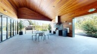 Located at 7900 Southeast Luther Road in Portland, Oregon, this single-story amenity clubhouse serves as a central gathering hub within a modern residential community. The building's architecture is defined by rich warm-toned cedar wood cladding and a distinctive vaulted wood-paneled ceiling that extends over a spacious covered outdoor pavilion, creating an inviting indoor-outdoor transition. Large floor-to-ceiling glass doors and windows allow abundant natural light to flow into the interior while offering clear sightlines to the surrounding grounds. The covered pavilion features polished concrete flooring and open roll-up garage-style access, seamlessly connecting interior and exterior spaces. Outdoor areas include a paved patio with Adirondack-style seating arranged around a central fire pit, ideal for communal gatherings. Lush green lawns, mature deciduous trees, and thoughtfully landscaped gravel beds frame the property. A secondary exterior structure features a clean, modern panel facade with large windows and an adjacent lounge chair patio. The broader community setting includes a multi-story residential building visible in the background, all surrounded by an open natural landscape with tall Pacific Northwest trees under wide Pacific Northwest skies.