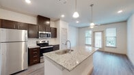 Located in Forney, TX, this single-floor residence offers a thoughtfully designed open-concept layout with modern finishes throughout. The kitchen serves as the centerpiece of the home, showcasing a large granite-topped island with an integrated sink, dark espresso cabinetry, a classic subway tile backsplash, and a full suite of stainless steel appliances including a refrigerator, range, and built-in microwave. Pendant lighting above the island adds a polished touch, while recessed lighting and crown molding elevate the overall aesthetic. Wood-look flooring flows seamlessly through the kitchen and living areas, creating a cohesive and easy-to-maintain surface. The living area is bright and airy, benefiting from natural light that pours in through large windows fitted with blinds, and a ceiling fan promotes comfortable airflow. The bedroom is generously sized with plush carpeting, dual windows offering neighborhood views, and its own ceiling fan. The bathroom is clean and functional, featuring granite countertops that echo the kitchen's design language, dark cabinetry, wood-look flooring, and a glass-enclosed walk-in shower with white tile surround. Together, these spaces form a cohesive, move-in-ready home with consistent, contemporary finishes.