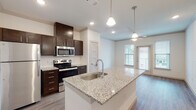 Located in Forney, TX, this single-floor residence offers a thoughtfully designed open-concept layout with modern finishes throughout. The kitchen serves as the centerpiece of the home, showcasing a large granite-topped island with an integrated sink, dark espresso cabinetry, a classic subway tile backsplash, and a full suite of stainless steel appliances including a refrigerator, range, and built-in microwave. Pendant lighting above the island adds a polished touch, while recessed lighting and crown molding elevate the overall aesthetic. Wood-look flooring flows seamlessly through the kitchen and living areas, creating a cohesive and easy-to-maintain surface. The living area is bright and airy, benefiting from natural light that pours in through large windows fitted with blinds, and a ceiling fan promotes comfortable airflow. The bedroom is generously sized with plush carpeting, dual windows offering neighborhood views, and its own ceiling fan. The bathroom is clean and functional, featuring granite countertops that echo the kitchen's design language, dark cabinetry, wood-look flooring, and a glass-enclosed walk-in shower with white tile surround. Together, these spaces form a cohesive, move-in-ready home with consistent, contemporary finishes.