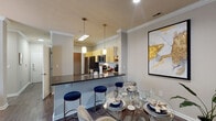 This thoughtfully designed single-floor residence offers a seamless blend of comfort and modern style throughout its well-proportioned layout. The living room serves as the heart of the home, featuring crown molding, dark hardwood-style flooring, and a large sliding glass door fitted with vertical blinds that bathe the space in natural light. The open-concept kitchen is equipped with light wood cabinetry, dark granite countertops, stainless steel appliances, and a breakfast bar with stylish pendant lighting overhead — ideal for casual dining or entertaining. Adjacent to the kitchen, the dedicated dining area accommodates a full-sized table and is accented by warm neutral tones and decorative wall art. A connecting hallway with crown molding and an arched architectural detail leads to the private quarters. The bedrooms are carpeted and well-lit by natural light from sizable windows, offering a calm and restful atmosphere. The bathroom features a granite-topped vanity with warm wood cabinetry, a tub-shower combination, and direct access to a spacious walk-in closet with wire shelving. Throughout the residence, consistent use of neutral tones, crown molding, and hardwood-style flooring creates a cohesive and inviting ambiance.