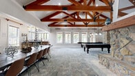 Located at 8940 N Congress Ave in Kansas City, Missouri, this expansive single-story facility offers a generously sized floor plan designed for community gatherings and multipurpose use. The architectural centerpiece is the dramatic vaulted ceiling supported by rich, exposed wood trusses and complemented by striking natural stone columns, creating a warm yet grand atmosphere throughout the main area.

The open main hall accommodates both a large dining table for group meals and a billiards table, with patterned carpet underfoot and abundant natural light streaming through multiple windows. A kitchenette features warm wood cabinetry, white quartz countertops, and a bold hexagonal tile backsplash. Adjacent to the main hall, a second, more intimate dining room offers a solid wood table with upholstered seating, ideal for private meetings or smaller gatherings.

The hallway entrance is finished with light stone tile flooring and features an elegant glass-panel barn door, leading visitors into the facility. A dedicated office provides two workstations with natural light. Additional spaces include a game room with poker tables and wall-mounted television, as well as arcade machines in a separate lounge area, rounding out a well-equipped, amenity-rich environment.