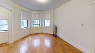 This single-floor studio offers a thoughtfully arranged layout that maximizes its compact footprint. The main living area is highlighted by a angled bay window configuration fitted with roller shades, allowing for controlled natural light while providing views of the surrounding urban streetscape. Warm-toned hardwood floors run throughout the main living space, lending a classic character to the interior. A cast-iron radiator positioned near the bay window provides traditional heating, a nod to the building's older architectural roots. The textured ceiling adds additional period detail to the space.

The separate kitchen features tile flooring, white cabinetry, a gas range, a full-size refrigerator, and a sink with countertop workspace. A doorway connects the kitchen directly to the main entry, creating a practical flow through the unit. An additional smaller room off the main living area offers flexible use as a sleeping nook or storage space.

The full bathroom includes a tub-shower combination with a curtain rod, a white vanity with under-sink storage, and blue ceramic tile accents along the walls, complemented by neutral tile flooring. Overall, the space reflects a functional, classic urban character.