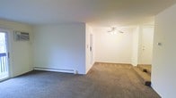 This single-floor space offers a clean, straightforward layout with a functional and understated design. The living area is finished with wall-to-wall carpeting in a neutral tone, providing a comfortable underfoot feel throughout the space. Crisp white walls create a bright, open atmosphere, and the room benefits from a ceiling fan that provides both light and air circulation. A baseboard heating system runs along the perimeter of the room, ensuring efficient climate control during cooler months. The open layout between the main living area and the adjoining room allows for a natural flow, making the space feel connected and versatile. A doorway visible in the interior wall suggests additional rooms beyond, contributing to the overall functional layout of the unit. The minimalist design and neutral color palette provide a blank canvas, making the space highly adaptable to a variety of furnishing and decorating styles. Overall, this space presents a practical and comfortable environment suited for everyday living.