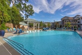 Located at 1185 Collier Rd NW in Atlanta, Georgia, this beautifully designed outdoor amenity area offers a resort-inspired experience for its residents. At the heart of the space is a generously sized swimming pool with crystal-clear blue water, complemented by a shallow tanning ledge outfitted with chaise loungers — perfect for relaxing in the Georgia sunshine. The pool deck is lined with additional lounge chairs and seating, providing ample space for sunbathing and socializing. A charming pergola structure offers a shaded retreat along one side of the pool, while a covered pavilion provides further shelter and gathering space. The adjacent clubhouse building features classic brick and white-siding architecture with a standing-seam metal roof, large French doors, and traditional wall-mounted lantern lighting that lend a timeless aesthetic to the overall setting. Beyond the pool, a paved brick courtyard hosts a circular stone fire pit surrounded by cushioned seating, nestled among mature trees and lush greenery that create a sense of privacy and tranquility. Stone retaining walls and well-maintained landscaping with native shrubs and seasonal plantings frame the entire outdoor area, adding natural texture and color. The space balances active recreation with quiet relaxation seamlessly.