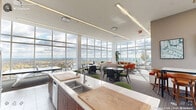 Located at 110 N Corcoran St in the heart of Durham, NC, this sophisticated clubroom offers a generously sized, single-floor amenity space designed for both social gatherings and relaxed lounging. The room is defined by dramatic floor-to-ceiling glass curtain walls that wrap around multiple sides, flooding the interior with abundant natural light and framing sweeping panoramic views of the Durham skyline and surrounding landscape.

The open-plan layout is organized into distinct functional zones. A well-appointed kitchen anchors one side of the space, featuring warm wood cabinetry, white quartz countertops, a large island with bar seating, stainless steel appliances, and a double sink — ideal for catering or casual entertaining. Adjacent to the kitchen, a bar-height counter with upholstered green stools provides an informal dining and socializing area.

The lounge zone is furnished with vibrant orange accent chairs, a neutral sofa, and a low coffee table, creating an inviting conversational setting bathed in sunlight. Multiple dining and meeting tables with mixed seating, including green-cushioned chairs, are distributed throughout the carpeted areas, offering flexible arrangements. A wall-mounted flat-screen TV and a linear fireplace add functional and atmospheric touches. Access to an outdoor terrace further extends the space, offering alfresco enjoyment with expansive open-air views.