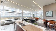 Located at 110 N Corcoran St in the heart of Durham, NC, this sophisticated clubroom offers a generously sized, single-floor amenity space designed for both social gatherings and relaxed lounging. The room is defined by dramatic floor-to-ceiling glass curtain walls that wrap around multiple sides, flooding the interior with abundant natural light and framing sweeping panoramic views of the Durham skyline and surrounding landscape.

The open-plan layout is organized into distinct functional zones. A well-appointed kitchen anchors one side of the space, featuring warm wood cabinetry, white quartz countertops, a large island with bar seating, stainless steel appliances, and a double sink — ideal for catering or casual entertaining. Adjacent to the kitchen, a bar-height counter with upholstered green stools provides an informal dining and socializing area.

The lounge zone is furnished with vibrant orange accent chairs, a neutral sofa, and a low coffee table, creating an inviting conversational setting bathed in sunlight. Multiple dining and meeting tables with mixed seating, including green-cushioned chairs, are distributed throughout the carpeted areas, offering flexible arrangements. A wall-mounted flat-screen TV and a linear fireplace add functional and atmospheric touches. Access to an outdoor terrace further extends the space, offering alfresco enjoyment with expansive open-air views.