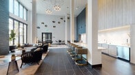 Located at 4949 Baltimore Dr in La Mesa, California, this elegantly designed single-floor clubhouse space showcases a seamless blend of modern architecture and upscale interior design. The main lounge commands attention with soaring double-height ceilings, dramatic geometric pendant lighting, and large floor-to-ceiling windows that flood the space with natural light. Underfoot, richly veined dark marble-style tile transitions to warm wood-look flooring, creating distinct zones within the open layout. A sleek built-in bar counter lined with upholstered swivel stools in brushed gold bases anchors one side of the room, complemented by custom wood cabinetry above. Curated seating arrangements featuring woven accent chairs, plush sofas, and stone-top coffee tables invite relaxed socializing throughout. Decorative sculptural wall art adds an artistic focal point, while a stacked dark stone accent wall with a recessed linear fireplace provides warmth and texture near the entry. A dedicated media lounge area features rich leather club chairs, ottomans, and a textured terrazzo-style stone feature wall with a wall-mounted display. A private conference room, also set against an expressive stone wall with light wood flooring, offers a functional yet refined setting for meetings. Indoor greenery and thoughtful lighting complete this polished, welcoming environment.