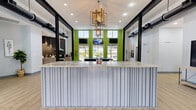 This contemporary single-floor community clubhouse offers a well-appointed, open layout designed for both social interaction and practical use. The space is defined by soaring double-height ceilings, bold black structural columns, and exposed ductwork that lend an industrial-chic character throughout.

The reception area serves as the focal point upon entry, featuring a sleek, ribbed-panel front desk topped with a marble-style surface, illuminated by a striking geometric gold chandelier. Vibrant lime-green accent walls and large windows frame views of the outdoors, flooding the space with natural light. Multiple wall-mounted TVs are positioned throughout for convenience and entertainment.

Adjacent to the reception, a dedicated workstation area offers a built-in counter with seating, a wall-mounted display, and a quiet nook for focused tasks.

The kitchen area is fully equipped with stainless steel appliances — including a range, refrigerator, and range hood — complemented by gray cabinetry, marble-look countertops, and a dramatic dark geometric tile backsplash. Pendant lighting adds a refined touch.

The lounge area is generously sized, featuring a pool table, comfortable seating arrangements, and direct access to the outdoors through large glass doors, making it an ideal gathering space for residents.